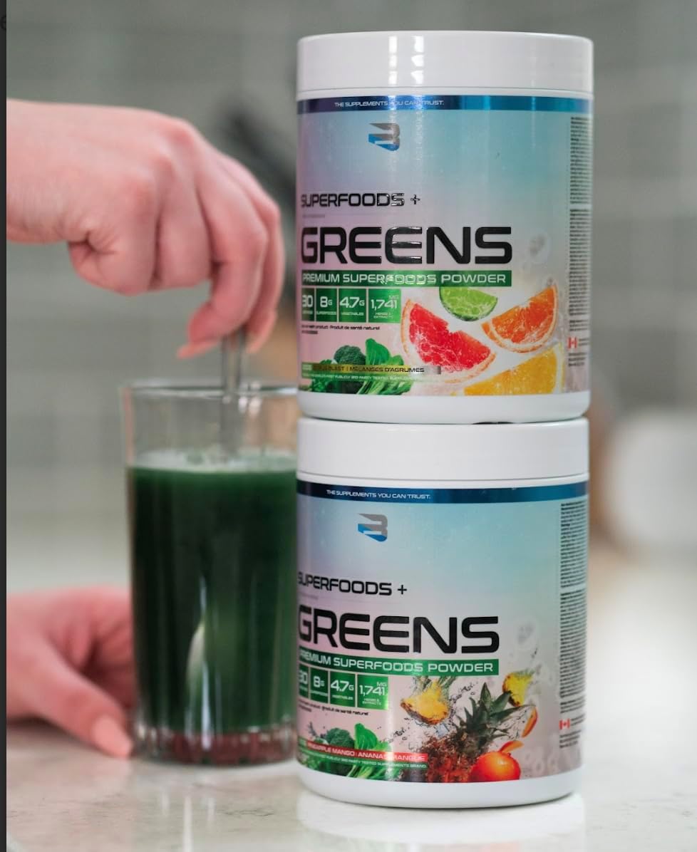 Two containers of SuperFoods + Greens powder with a glass of green juice on a countertop.
